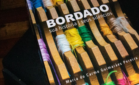 Fotografia do livro Bordado: sua história; seus silêncios. Na capa, várias plaquetas de madeira de enrolar linhas e linhas coloridas.